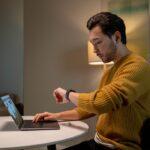 A man in a mustard sweater sits at a table, checking his smartwatch while working on a laptop.