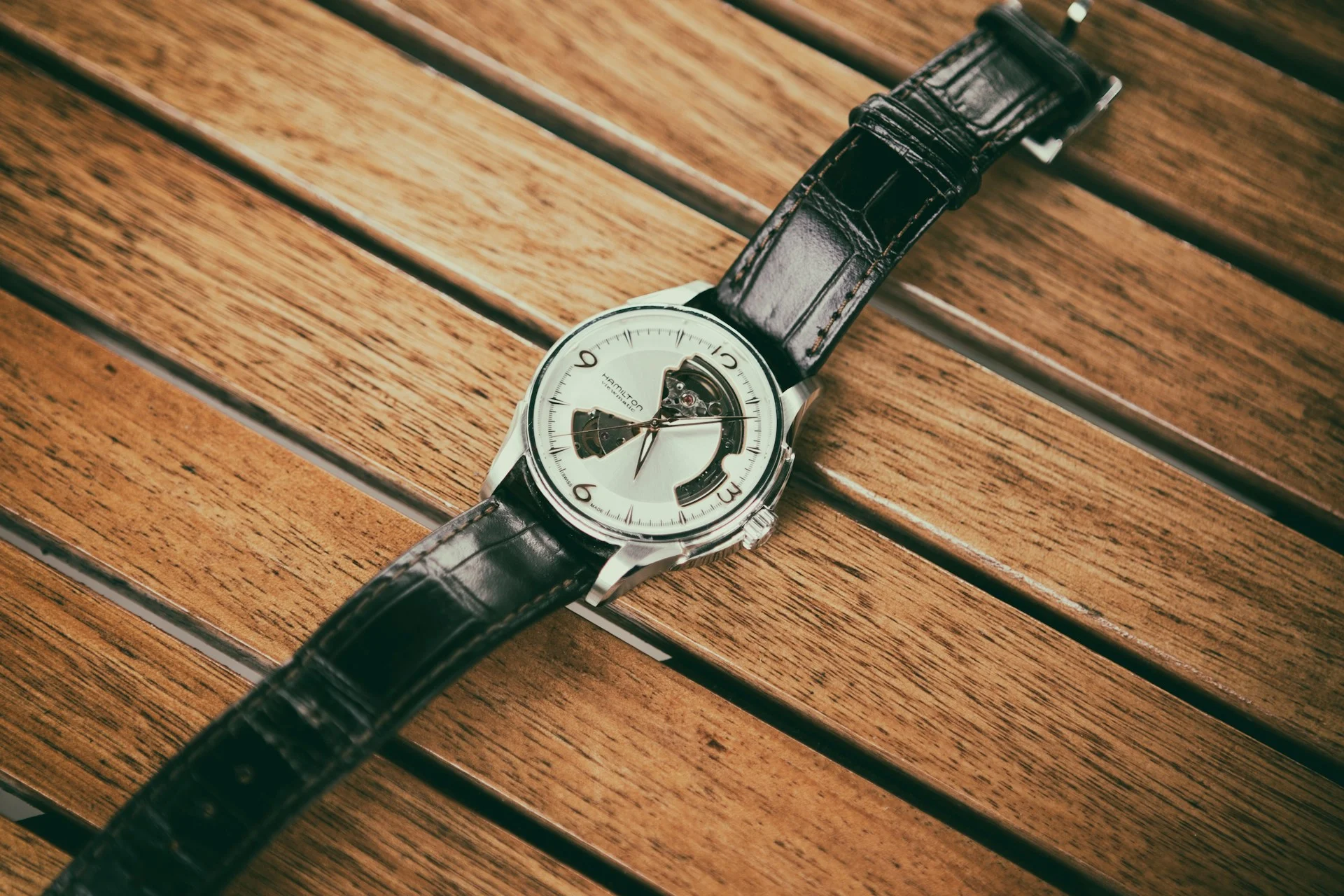A Hamilton open-heart watch with a black leather strap and a silver dial featuring exposed gears, resting on a wooden surface. 