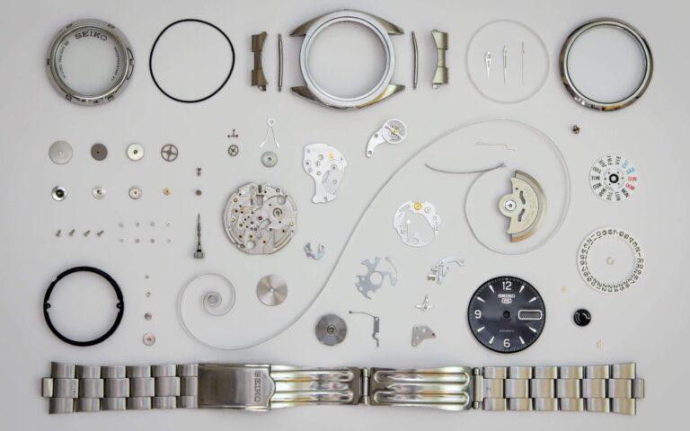 An array of disassembled wristwatch parts laid out neatly on a white background, including a watch face, various gears, springs, screws, and a metal band.