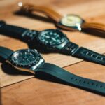 Three wristwatches with different watch materials: black rubber and steel, plastic, and brown leather with light dial on a wooden surface.