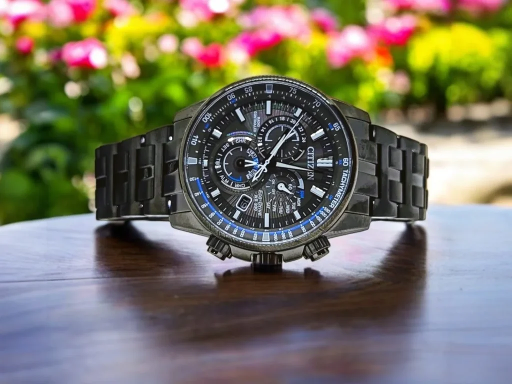 Citizen PCAT CB5887-55H sits on a wooden surface. The background is blurred with vibrant green and pink flowers.