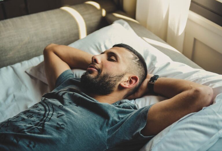A man with short dark hair and a beard lies on a bed, arms behind his head, wearing a smartwatch on his wrist to track his sleep.