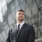A man in a black business suit and tie stands confidently in front of a modern glass building.