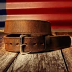 A brown full-grain leather belt with a silver buckle lies coiled on a wooden surface.