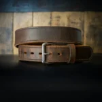 A brown leather belt with a silver buckle is coiled on a dark surface. The background features rustic wooden planks, giving a vintage feel.