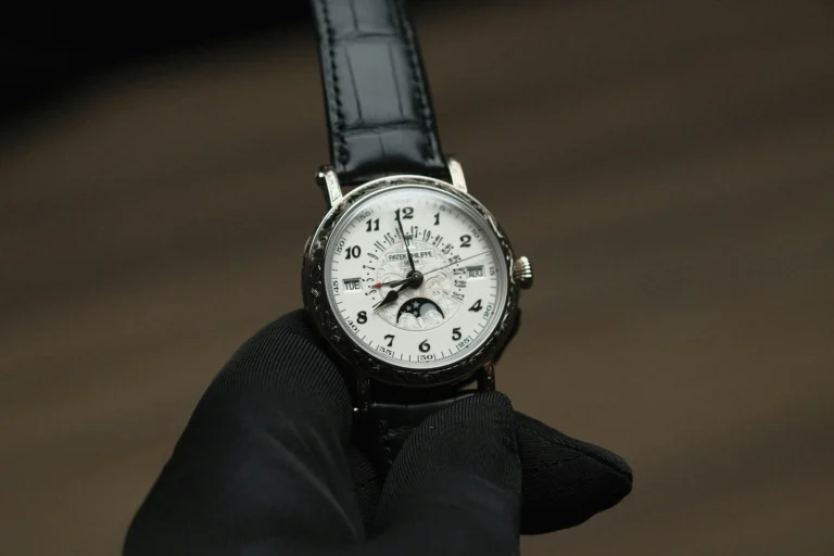 A gloved hand holding a luxury watch with a white dial, black numerals, and moon phase display, reflecting elegance and precision against a blurred brown background.