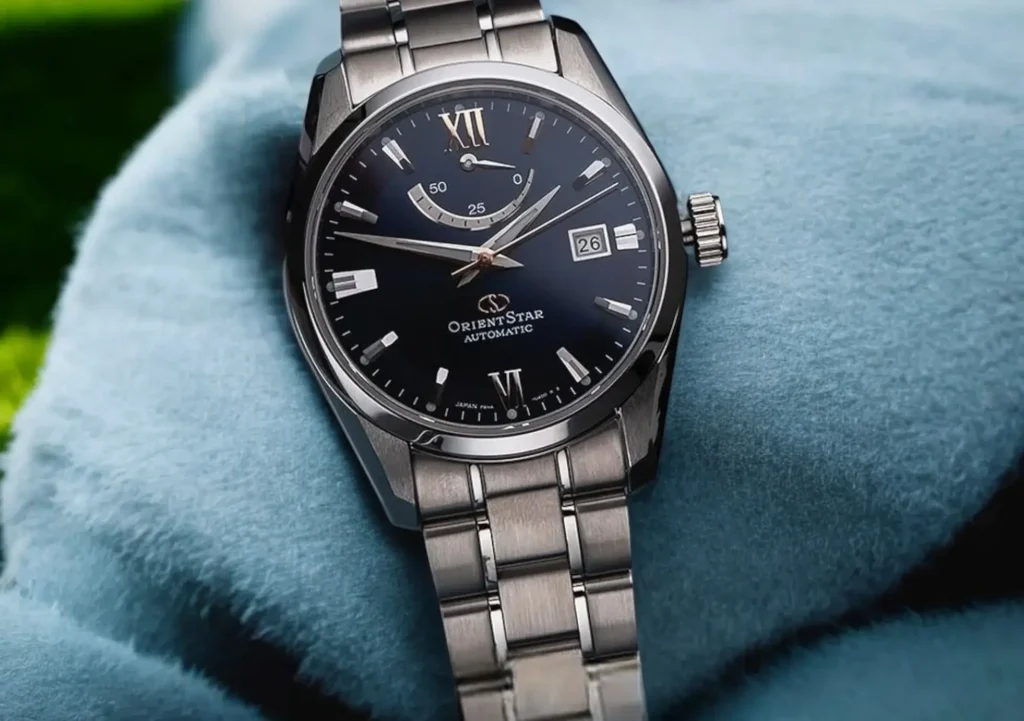 Orient Star stainless steel dress watch displayed on light teal-blue fabric with a dark navy blue dial in a modern, sophisticated setting.
