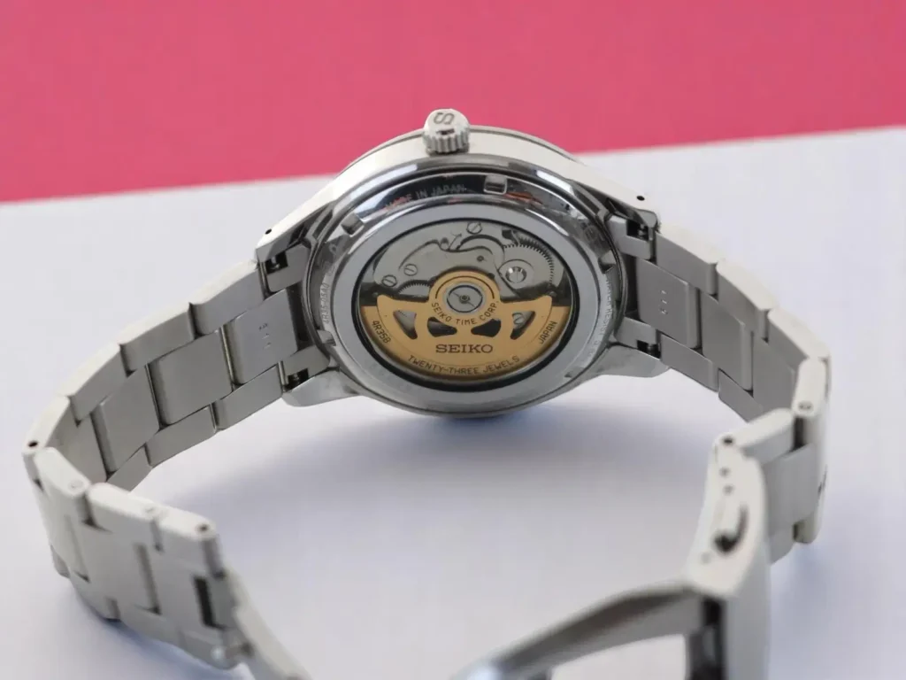 Seiko Presage Style60's SRPG05 automatic watch showcasing its exhibition case back and stainless steel bracelet on a white & pink surface.