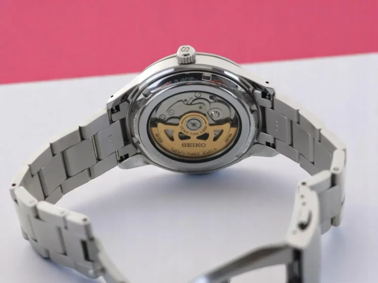 Seiko Presage Style60's SRPG05 automatic watch showcasing its exhibition case back and stainless steel bracelet on a white & pink surface.