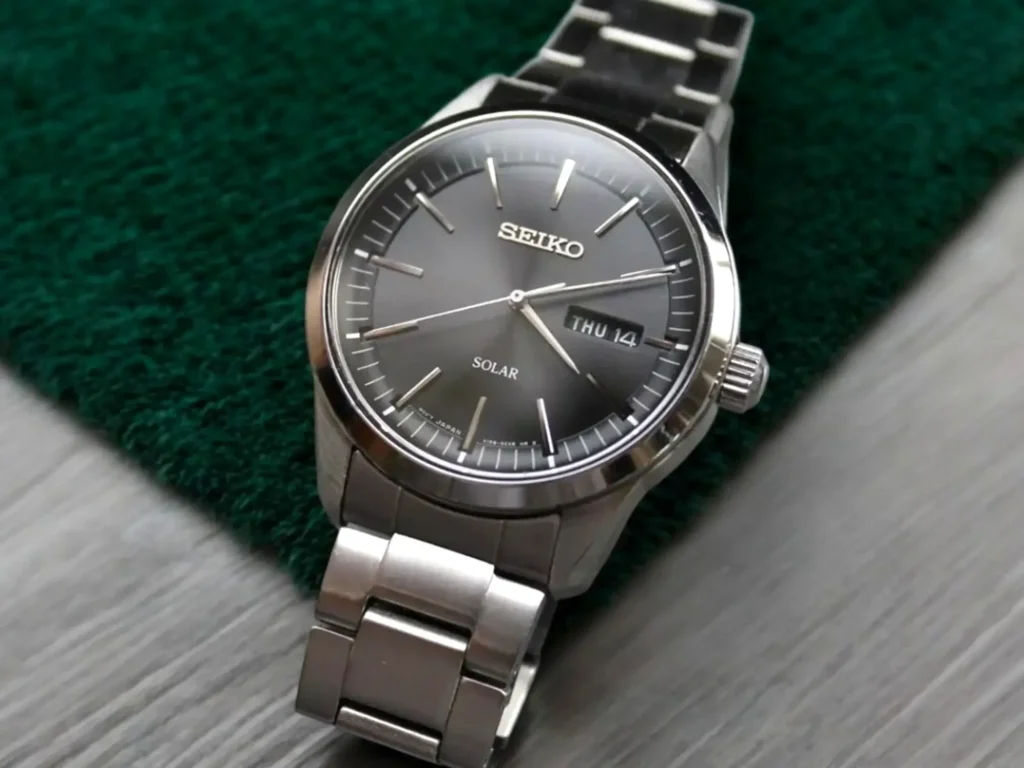 Close-up of the Seiko Solar SNE527 dress watch placed on a light gray wooden surface with dark teal-green plush fabric in the background.
