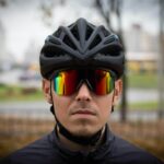 A cyclist wears a black helmet and reflective sports sunglasses with a serious expression, set against a blurred outdoor city background.