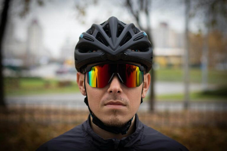 A cyclist wears a black helmet and reflective sports sunglasses with a serious expression, set against a blurred outdoor city background.
