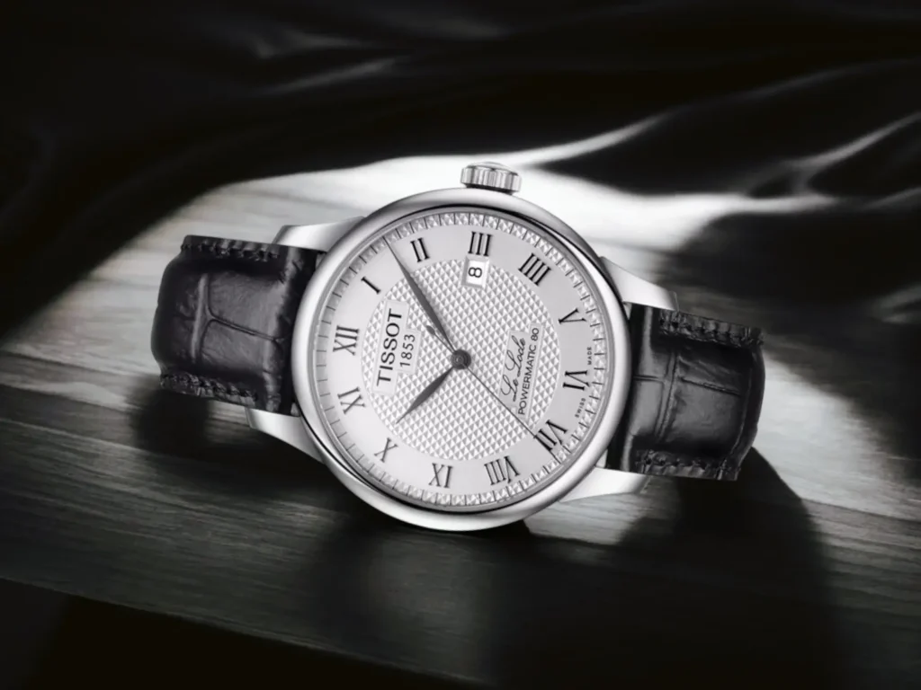 Tissot Le Locle Powermatic 80 dress watch with black leather strap displayed on a light gray wooden surface with dark gray fabric background in an elegant, dramatic setting.
