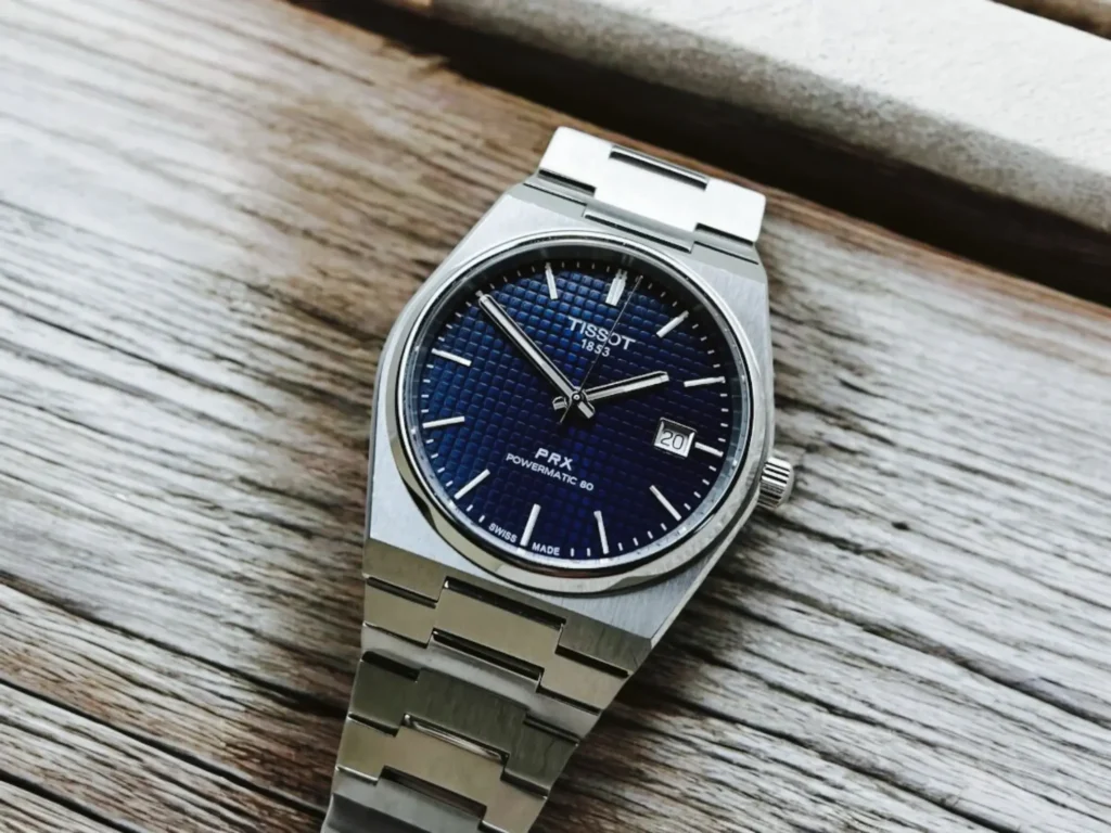 Tissot PRX Powermatic 80 dress watch placed diagonally on a light brownish-gray wooden surface with visible wood grain in a classic, minimalist setting.