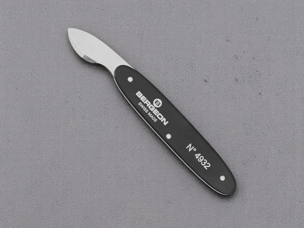 A small, curved stainless steel watch back removal tool with a black handle, labeled "Bergeon Swiss Made N° 4932," resting on a gray textured surface.