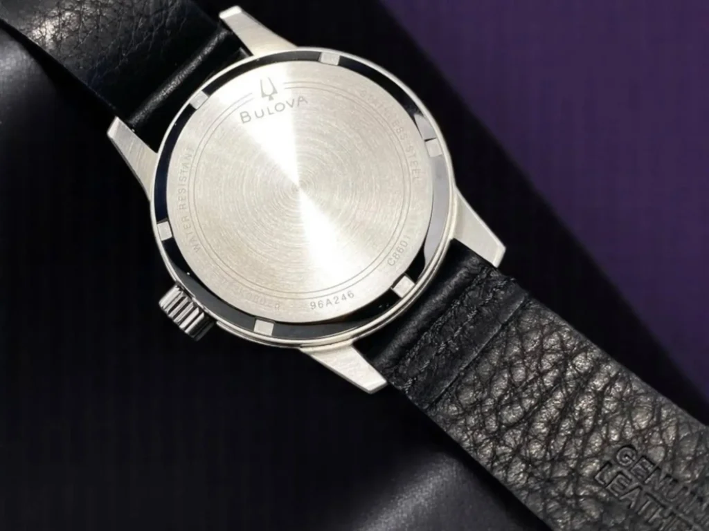 Back view of the Bulova Hack Watch 96A246 showing its solid screw-down caseback and black genuine leather strap, resting on black textured leather with a dark purple background.