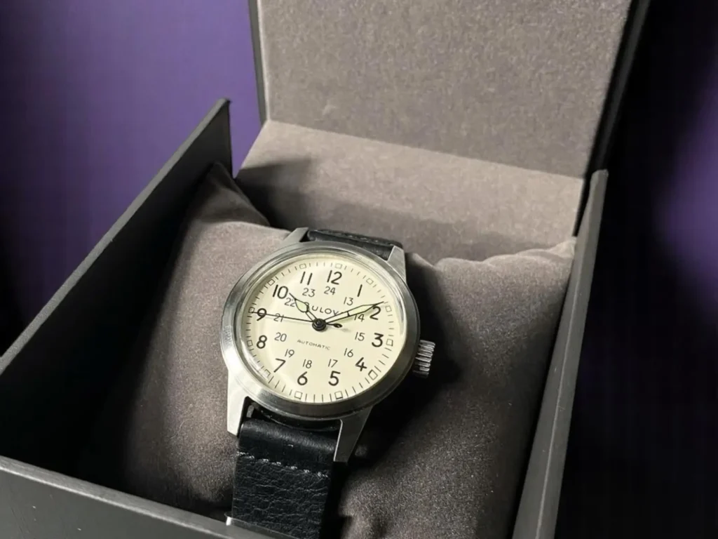 Bulova Hack Watch 96A246 sits on a gray cushion inside an open black box. The background is a deep purple.