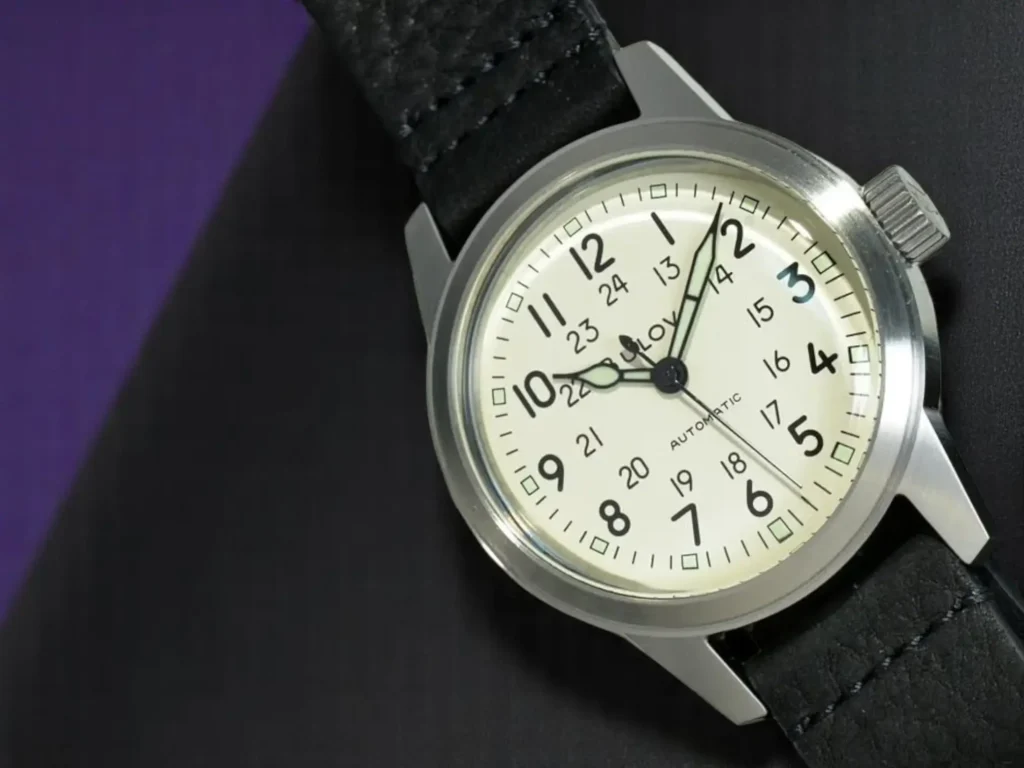 Bulova Hack Watch 96A246 with an oversized crown, photographed at a slight angle on black textured leather with a dark purple background.