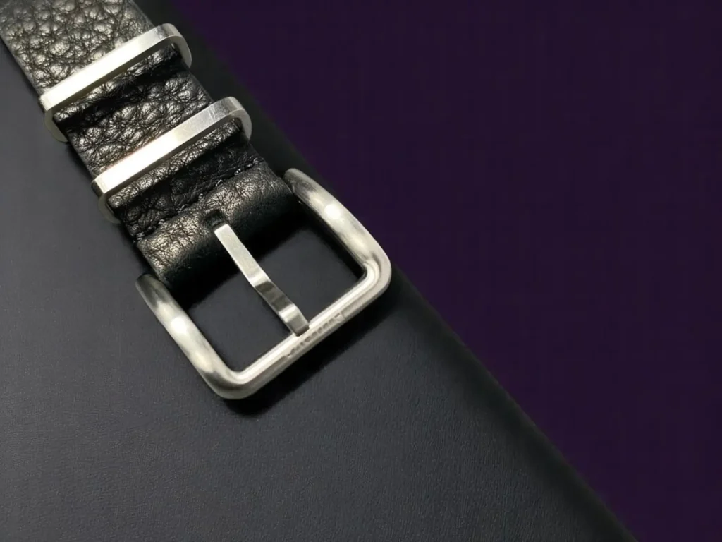 Close-up of the silver buckle on the black leather strap of the Bulova Hack Watch 96A246, resting on black textured leather with a dark purple background.