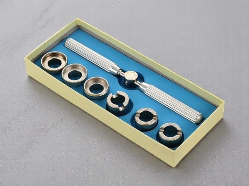 A boxed set featuring a silver Rolex-style case back removal tool with a textured handle and six dies. The entire box, resting against a gray surface, has a blue interior. 