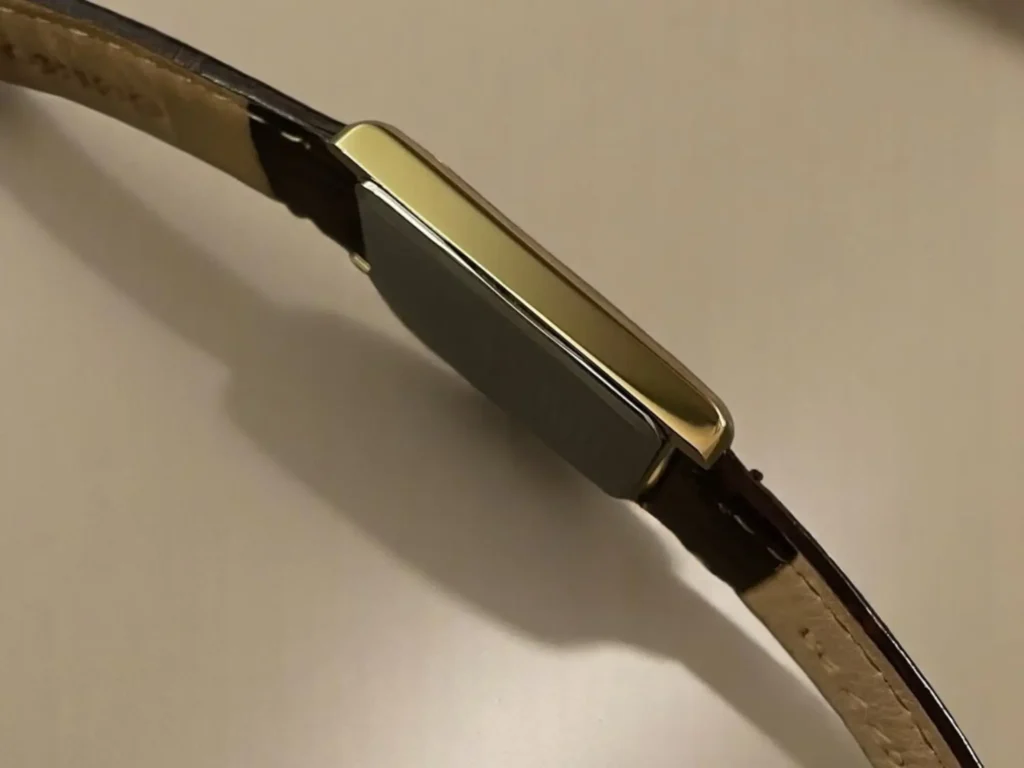 A side view of the Seiko Essentials watch featuring a gold-toned rectangular case and a dark brown leather strap, placed on a soft beige surface. 