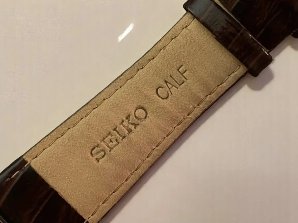 Close-up of a beige leather watch strap with "SEIKO" and "CALF" embossed on it. The strap has dark brown edges and visible stitching.