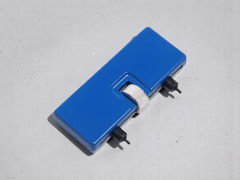 A blue rectangular watch back removal wrench with a silver knob and two black prongs rests on a light gray surface.