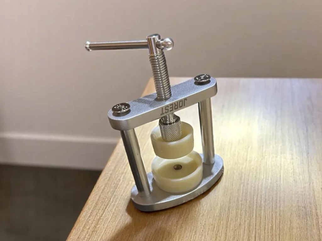 A metal watch press tool with a handle and cream-colored discs sits on a wooden table.