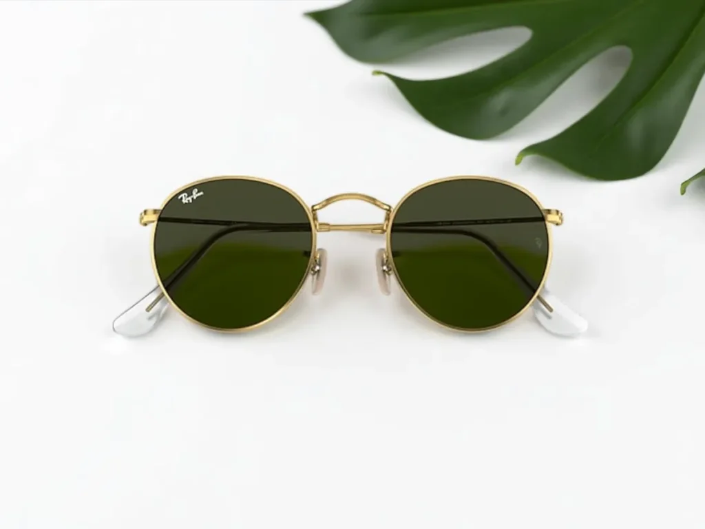 Ray-Ban golden round sunglasses with dark lenses on a white surface, accompanied by a large green leaf on the top right.