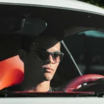 A young man wearing sunglasses sits in a car with red leather interior, partially shaded by sunlight.