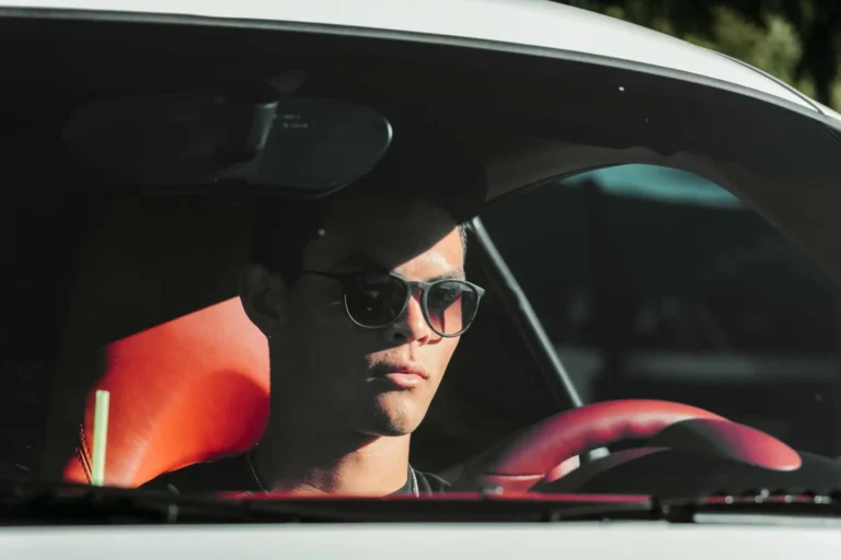 A young man wearing sunglasses sits in a car with red leather interior, partially shaded by sunlight.
