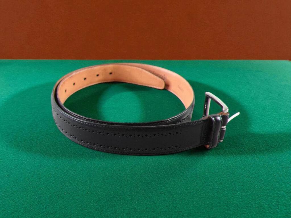 Allen Edmonds Manistee leather dress belt with a silver buckle is coiled on a green surface, contrasting against an orange-brown wall.