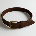 A brown casual leather belt with a bronze buckle is coiled on a light gray surface, casting a soft shadow.