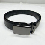 Mission Belt black leather dress belt with a smooth, rectangular metal buckle is coiled on a white surface.