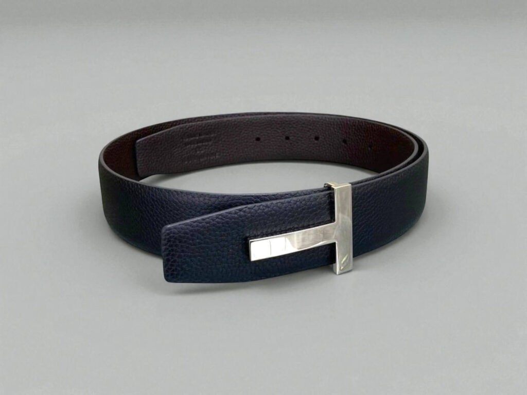 Tom Ford leather belt with a silver 'T' buckle, coiled neatly against a soft gray background.