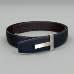 Tom Ford leather belt with a silver 'T' buckle, coiled neatly against a soft gray background.
