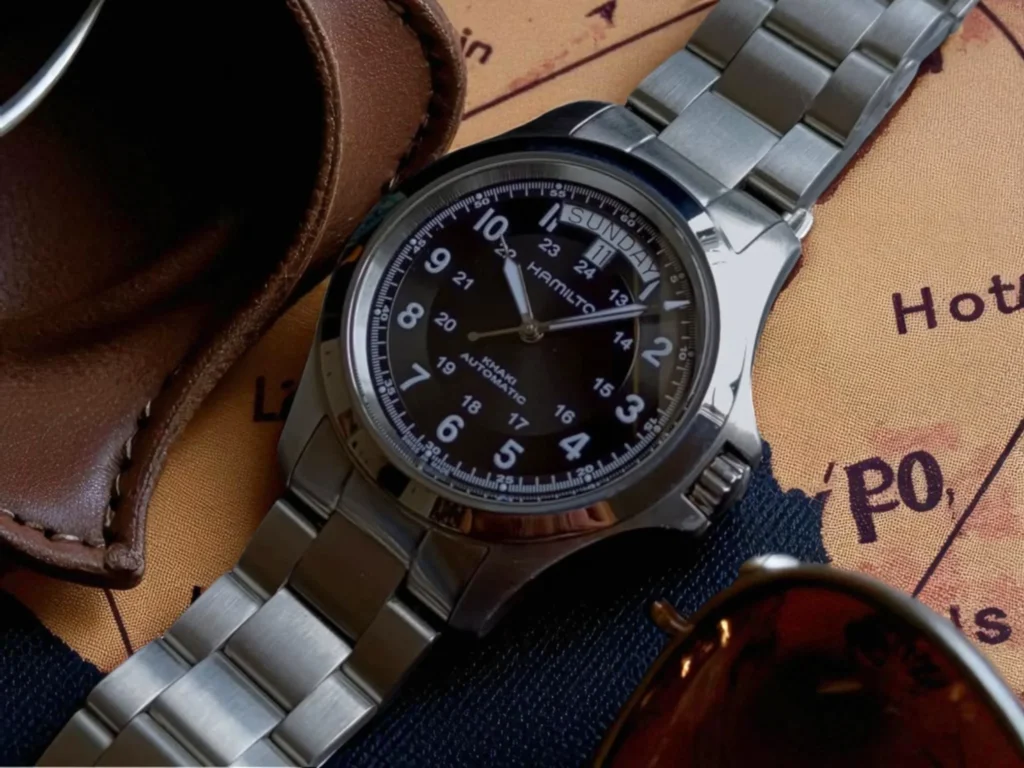Hamilton Khaki Field King Auto resting on a vintage map beside a brown leather case and sunglasses. 