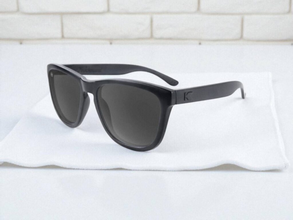 Knockaround Premiums polarized sunglasses with dark lenses rest on a white cloth. A brick wall acts as a soft background.