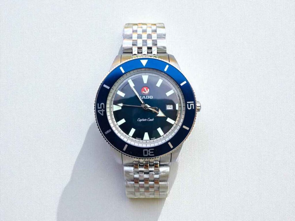 A silver Rado Captain Cook watch with a blue bezel, silver markers, and a stainless steel bracelet on a light background, casting a shadow.