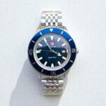 A silver Rado Captain Cook watch with a blue bezel, silver markers, and a stainless steel bracelet on a light background, casting a shadow.