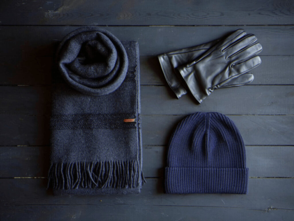 Dark cozy winter accessories on a wooden surface: a folded wool scarf, leather gloves, and a ribbed knit beanie.