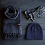 Dark cozy winter accessories on a wooden surface: a folded wool scarf, leather gloves, and a ribbed knit beanie.