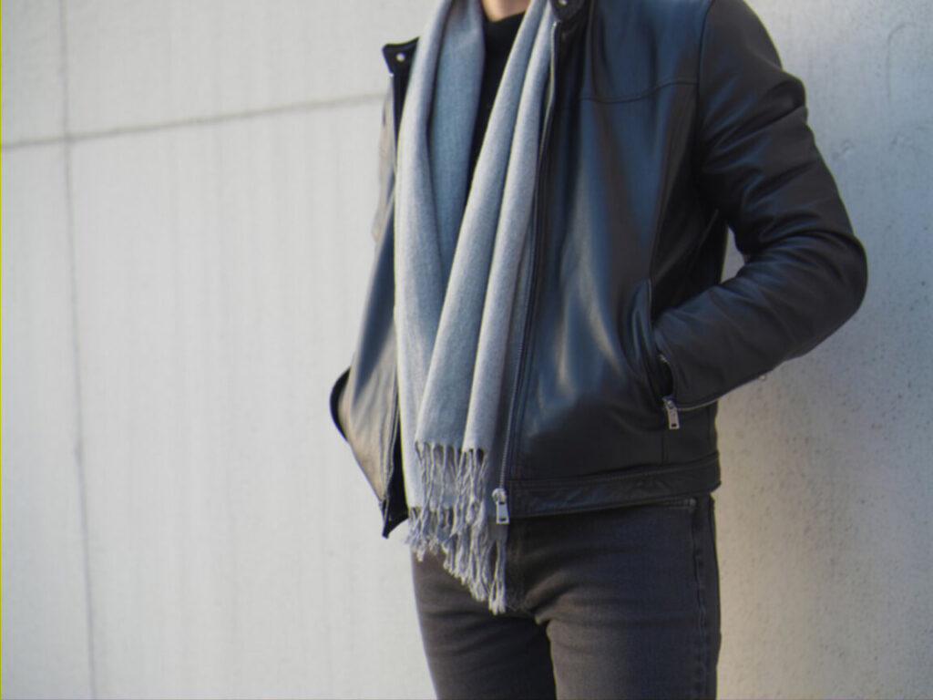A person wearing a black leather jacket and a light gray scarf, hands in pockets, leans against a light-colored wall.