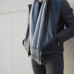 A person wearing a black leather jacket and a light gray scarf, hands in pockets, leans against a light-colored wall.
