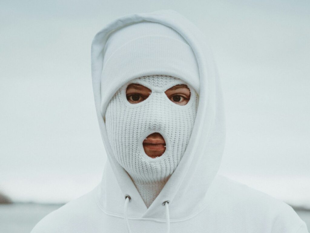 A person wearing a Balaclava hat and a hooded sweatshirt stands in a neutral outdoor setting.