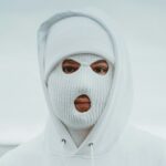 A person wearing a Balaclava hat and a hooded sweatshirt stands in a neutral outdoor setting.
