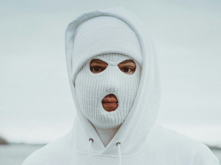A person wearing a Balaclava hat and a hooded sweatshirt stands in a neutral outdoor setting.