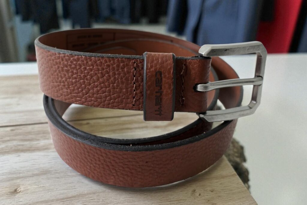 A brown Carhartt leather belt with a textured surface and a silver buckle is coiled on a wooden table. Clothing hangs in the blurred background.