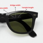 Close-up of black sunglasses highlighting lens width, bridge width, and temple length with arrows and labels. Sunglasses have dark green lenses.