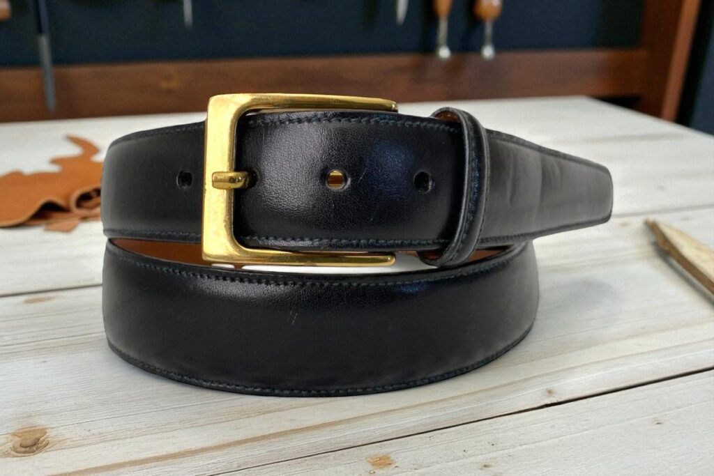 A black Trafalgar leather belt with a shiny gold buckle is coiled on a light wooden table. 